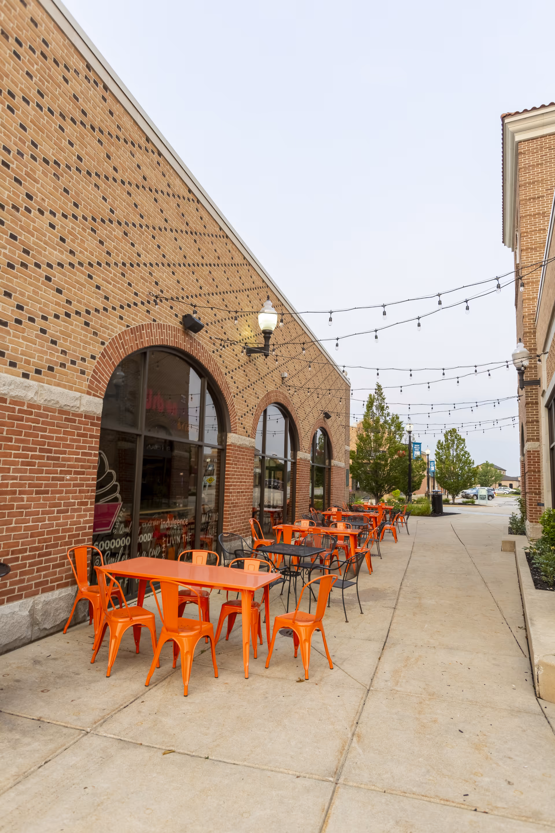 Outdoor café seating with bright orange tables and chairs along a brick building under string lights.