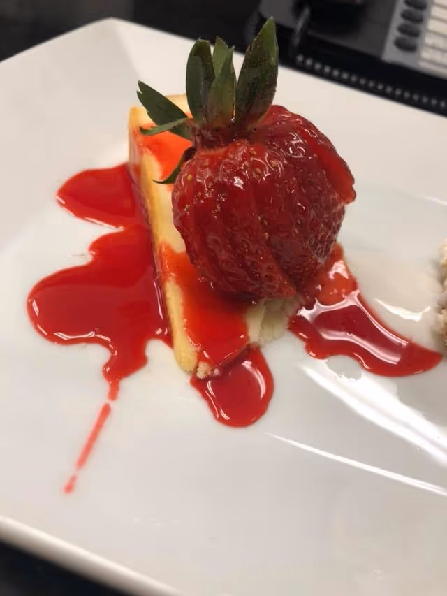 A slice of cheesecake topped with a whole strawberry and drizzled with red strawberry sauce on a white square plate.