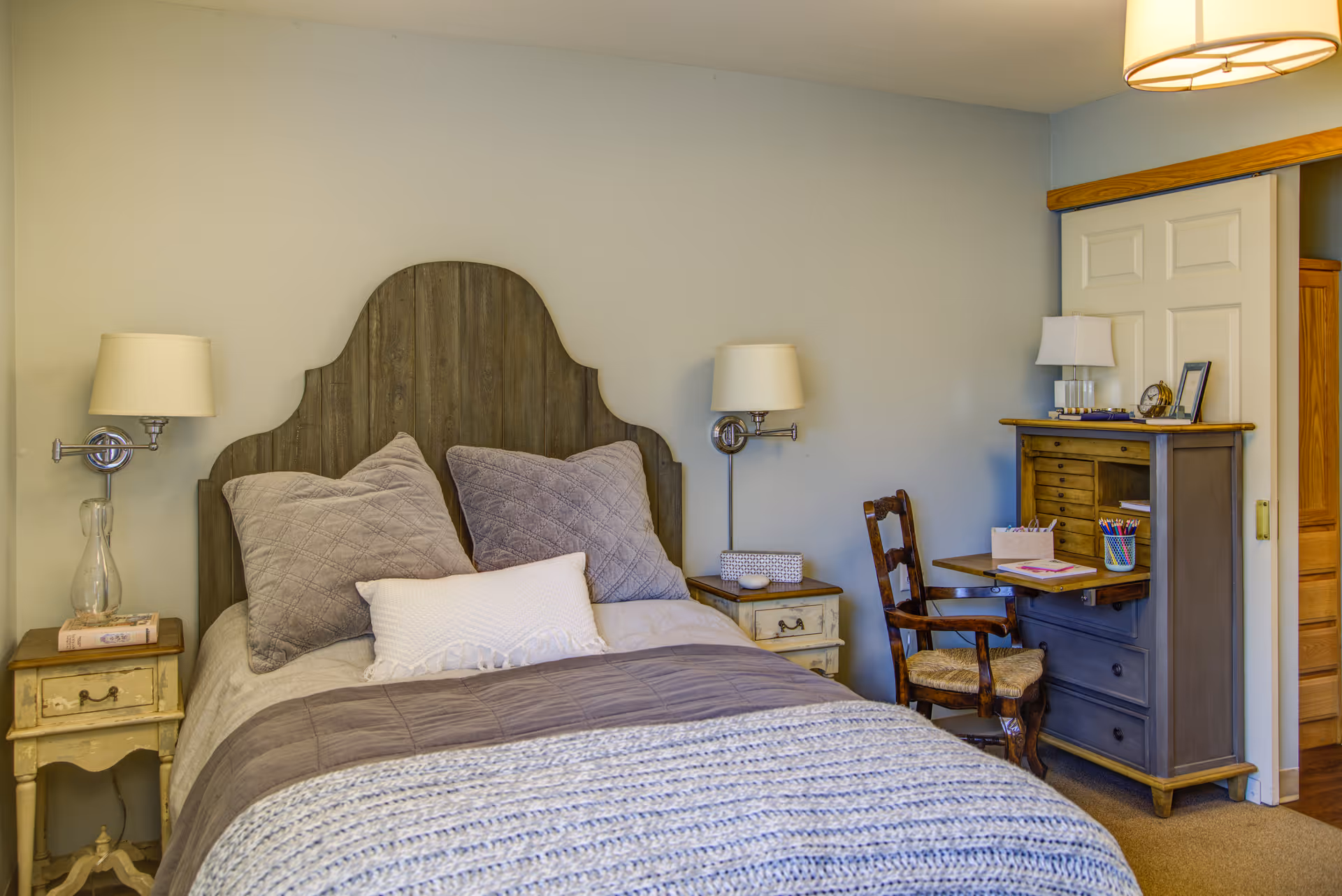 A cozy bedroom with a wooden headboard bed made with gray and white pillows and blankets. There are two bedside tables with lamps on either side of the bed. To the right, there is a wooden chair and a gray cabinet with a fold-down desk surface, holding some stationery and a lamp. The room has light-colored walls and carpeted floor.