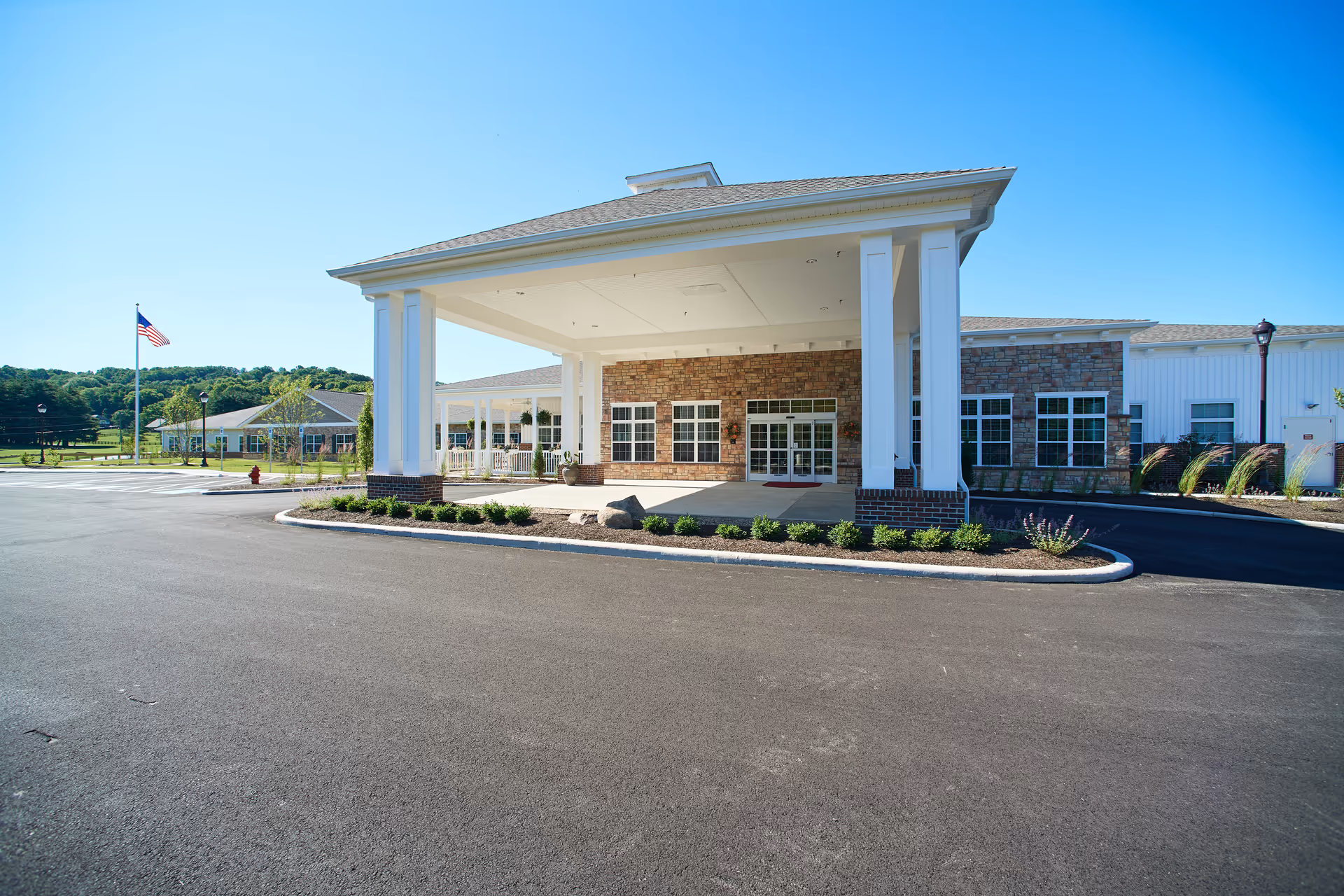 Front entrance of a single-story senior living facility with a covered porte-cochere, brick facade, and an American flag on a flagpole.