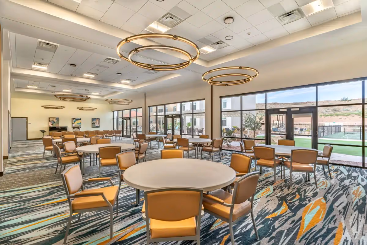 A spacious, well-lit dining room with multiple round tables surrounded by tan chairs. Large windows on one side provide a view of an outdoor area with greenery and a tennis court. The ceiling features modern circular light fixtures, and the floor is covered with a patterned carpet in shades of blue, gray, and orange.