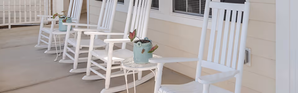 A row of white wooden rocking chairs lined up on a covered porch with small round tables between some of the chairs. Each table has a small decorative plant in a blue pot. The porch has beige siding and windows with blinds.