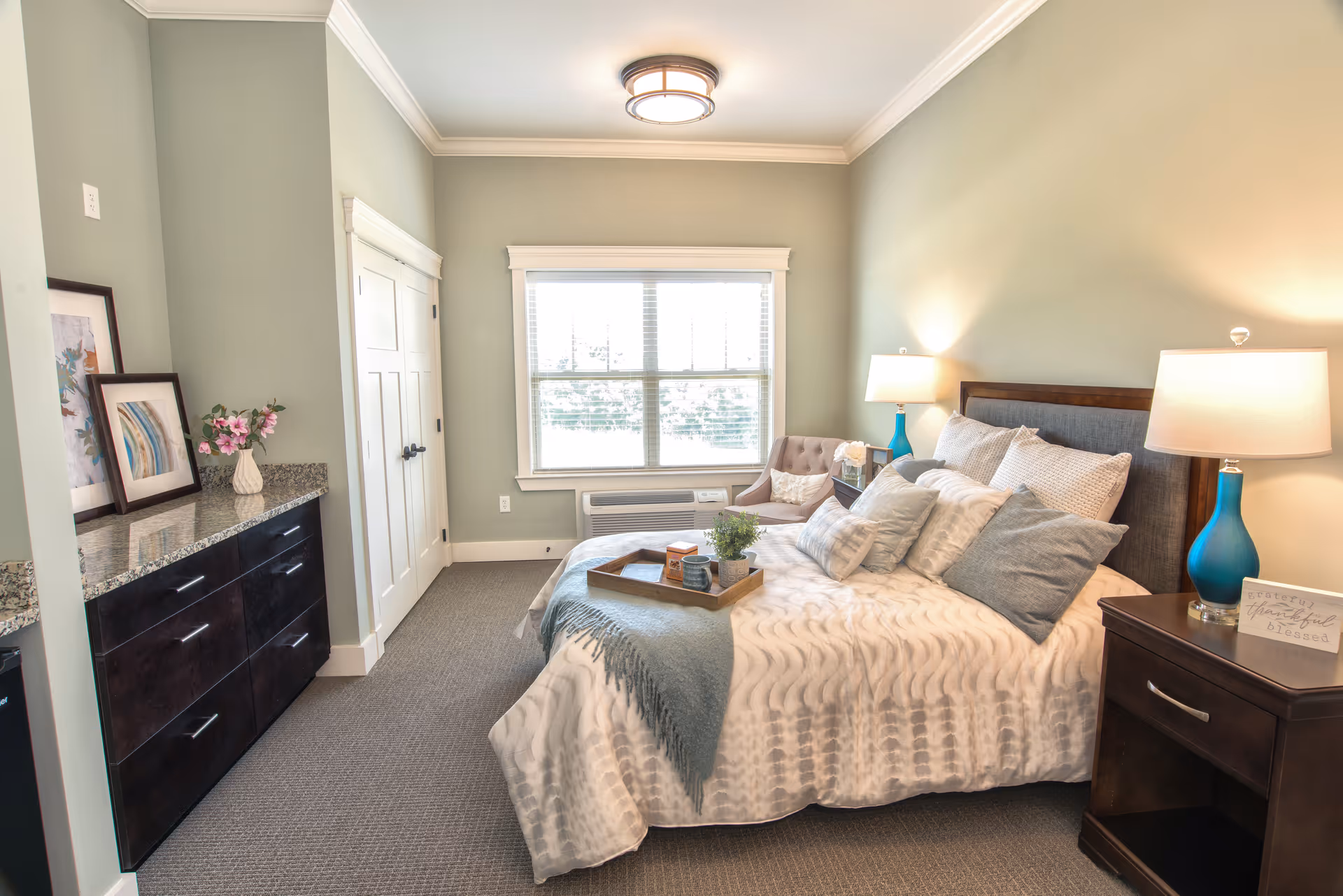 A cozy bedroom with a large bed covered in white and gray bedding and multiple pillows. There are two bedside tables with blue lamps and a small decorative sign. A window with white blinds lets in natural light. A cushioned chair is placed near the window. On the left side, there is a dark wood dresser with a granite countertop, decorated with framed pictures and a vase with pink flowers. The walls are painted light green and the floor is carpeted in gray.