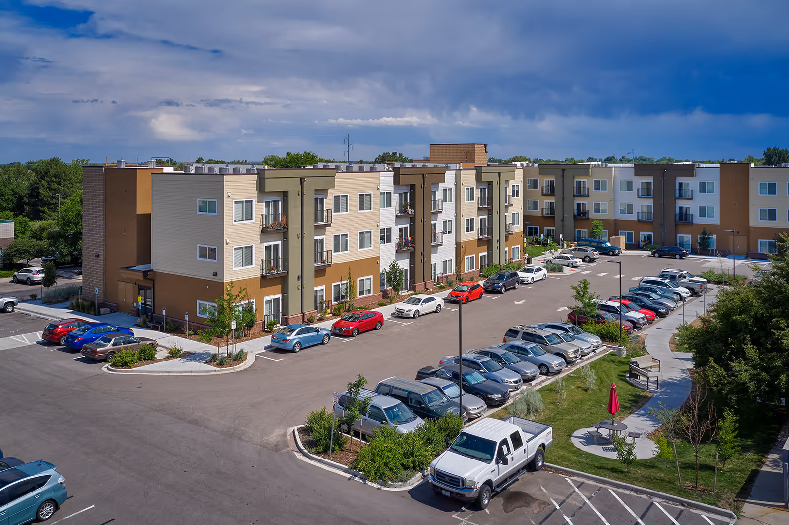 Exterior view of a three-story modern senior living building with parked cars, landscaped grounds, and a cloudy sky.