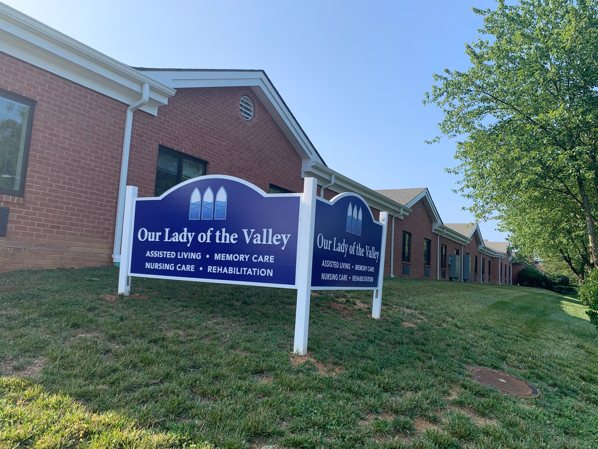Exterior view of a brick building with a blue sign in front that reads 'Our Lady of the Valley Assisted Living, Memory Care, Nursing Care, Rehabilitation' on a grassy area with a tree and clear sky.