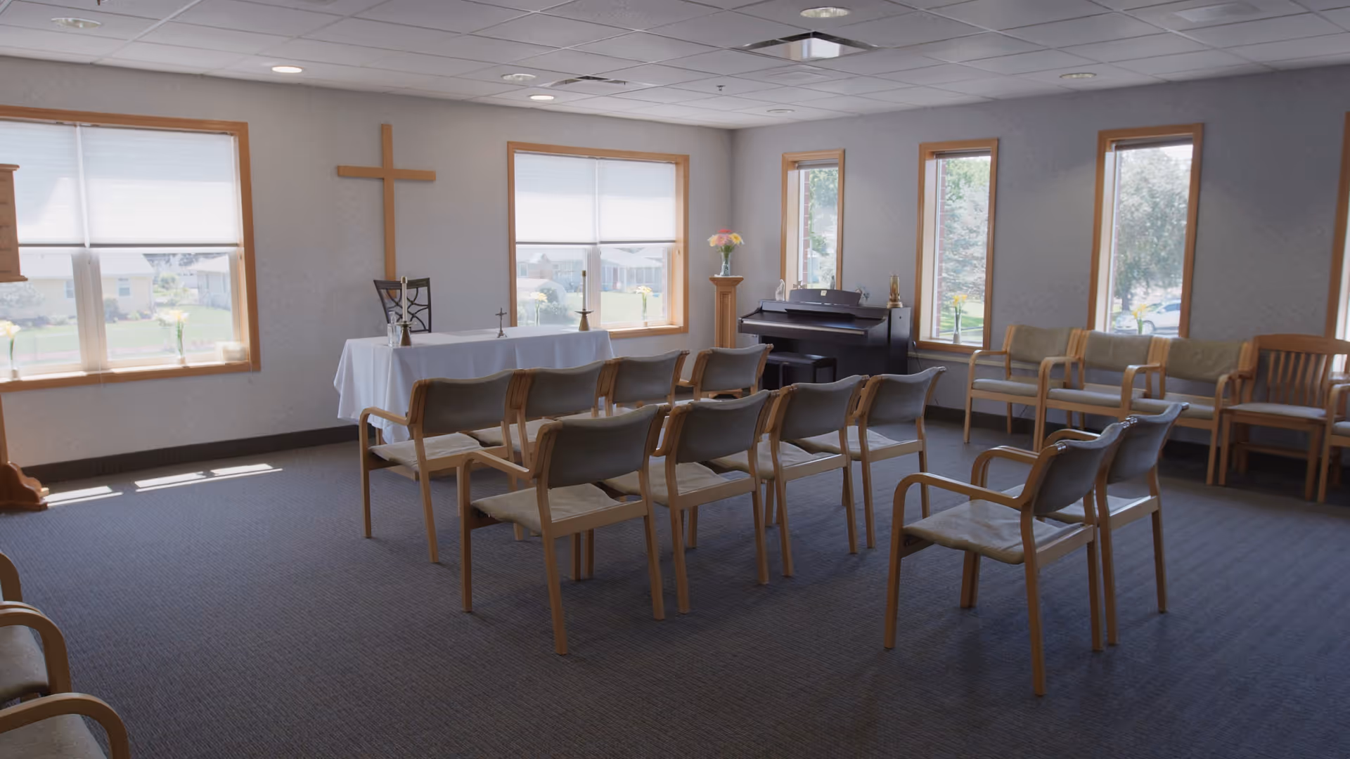 A small chapel or prayer room with rows of wooden chairs facing a table covered with a white cloth. A wooden cross is mounted on the wall behind the table. There is a piano in the corner and several windows letting in natural light. The room has a calm and serene atmosphere.