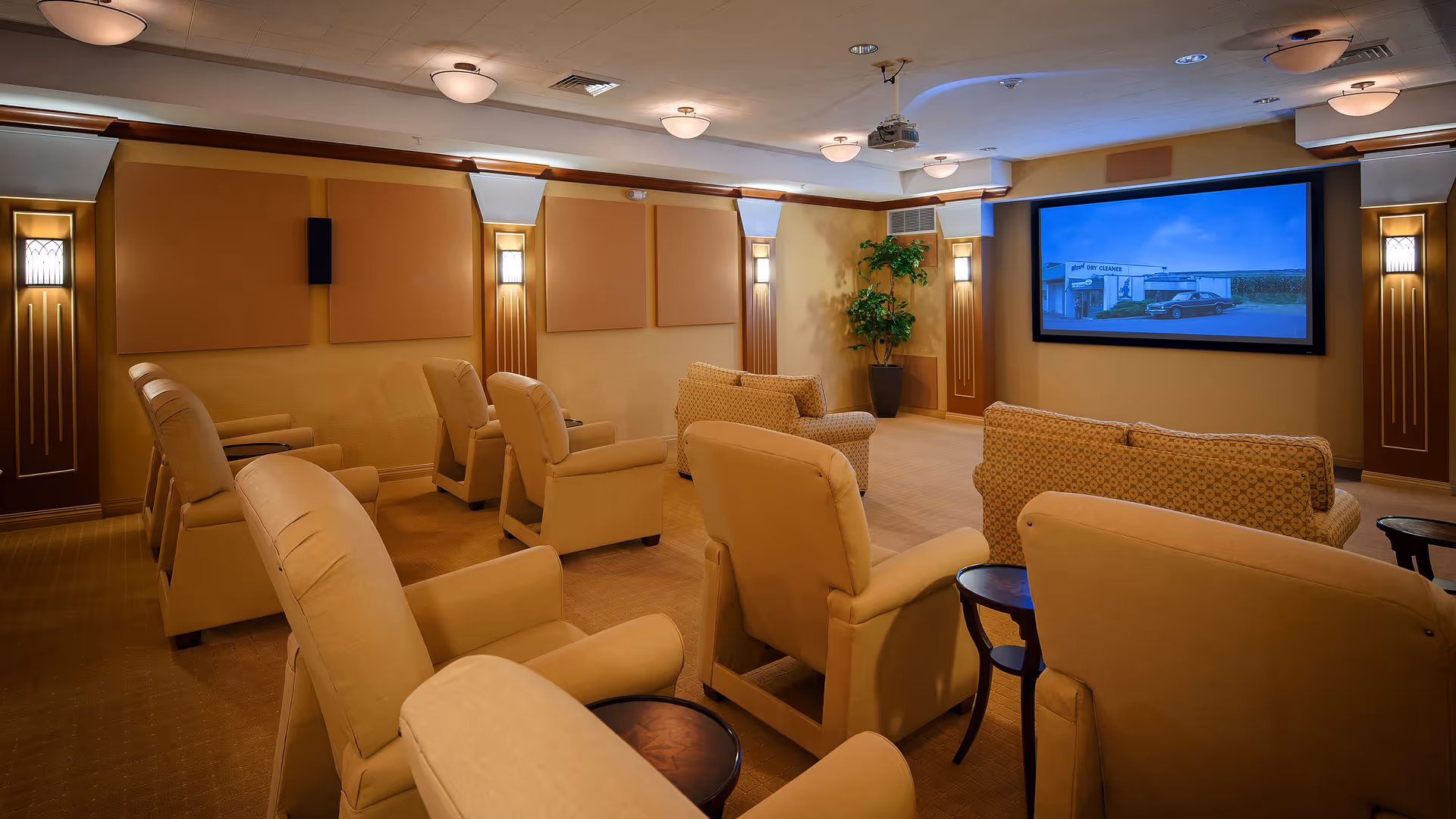 A cozy home theater room with multiple beige armchairs and sofas arranged facing a large screen on the wall. The room has warm lighting with wall sconces and a potted plant in the corner. The screen displays an image of a building and a car.