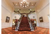 A grand interior staircase with a red carpet runner and floral decorations on the banisters, leading up to a balcony area. The room features a chandelier hanging from the ceiling, white paneled walls, and patterned carpet flooring. There are framed pictures on the walls and chairs placed near the staircase.