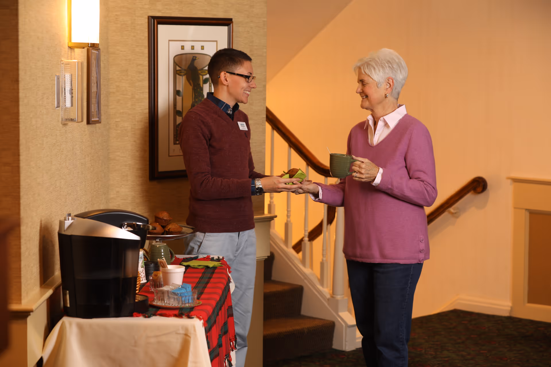 A young man wearing glasses and a maroon sweater hands a plate of food to an older woman with short white hair wearing a purple sweater, who is holding a green mug. They are standing in a warmly lit hallway near a staircase, with a table covered in a red and green cloth holding a coffee maker and snacks.