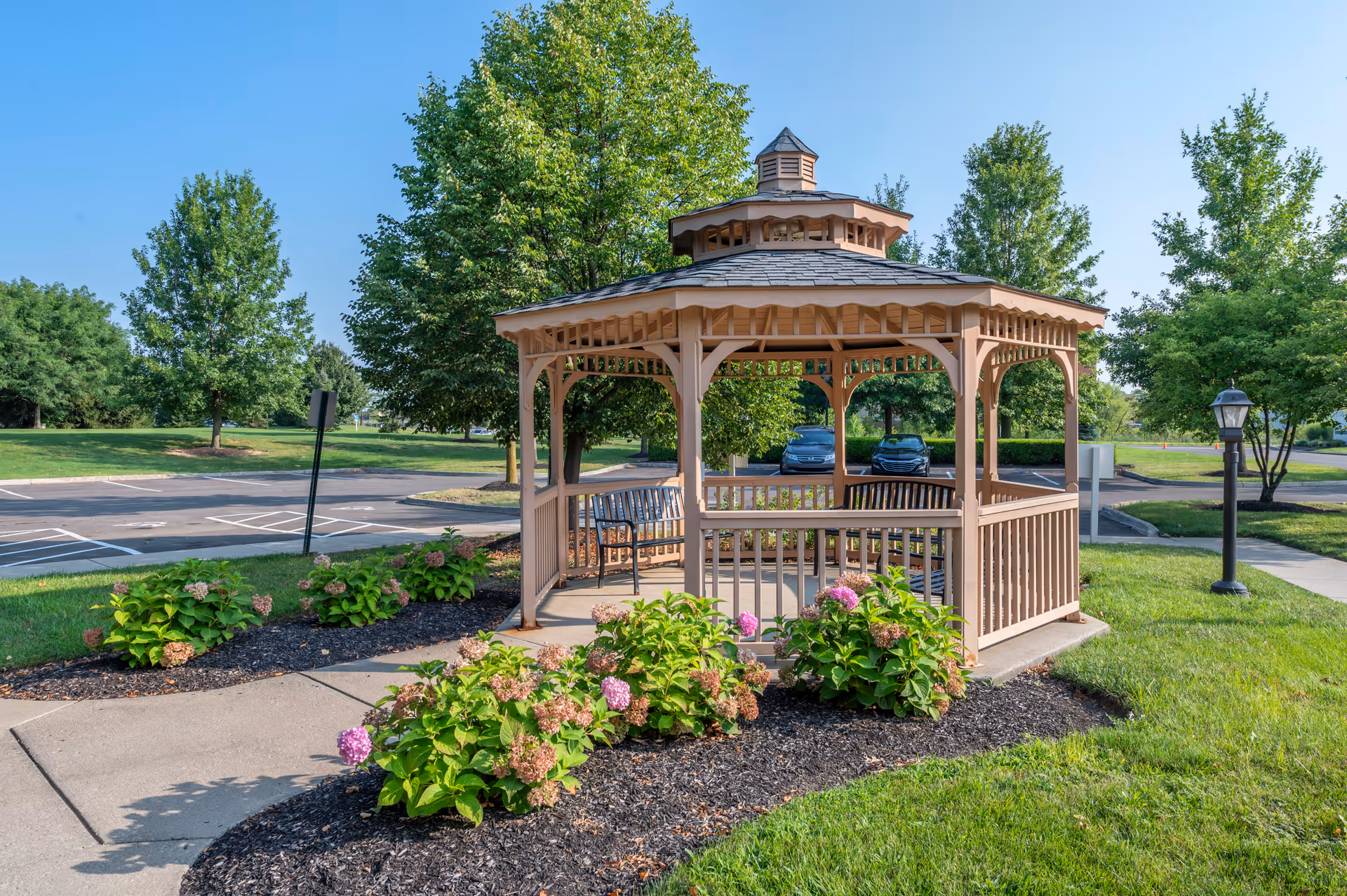 A wooden gazebo with benches inside, surrounded by green grass, flowering bushes, and trees. There is a paved walkway leading to the gazebo and a parking lot with cars in the background under a clear blue sky.