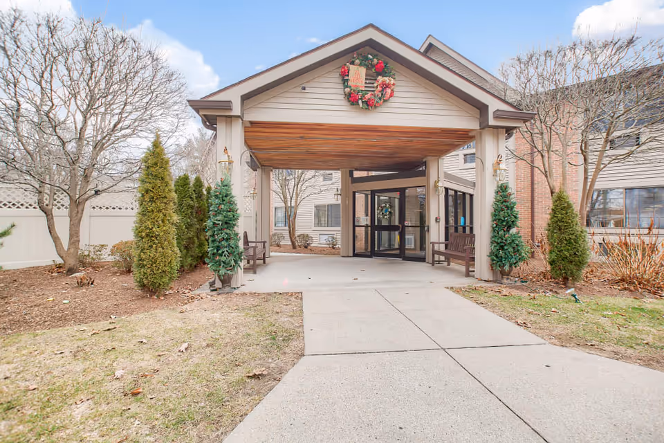 Entrance to a senior living facility with a covered driveway. The building has beige siding and brick accents. There are small evergreen trees and leafless deciduous trees on either side of the walkway. A festive wreath is hanging above the entrance.
