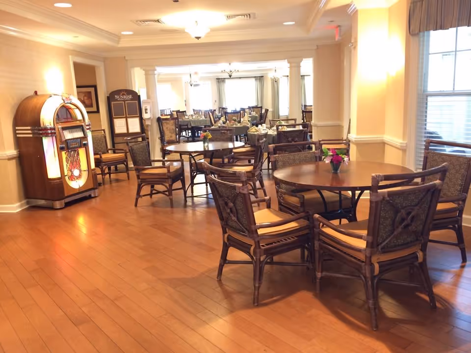 A spacious dining room with wooden floors and multiple round tables surrounded by cushioned chairs. A vintage jukebox is positioned against the left wall, and there is a signboard labeled 'Sunrise' near the jukebox. The room is well-lit with natural light coming through large windows on the right side.