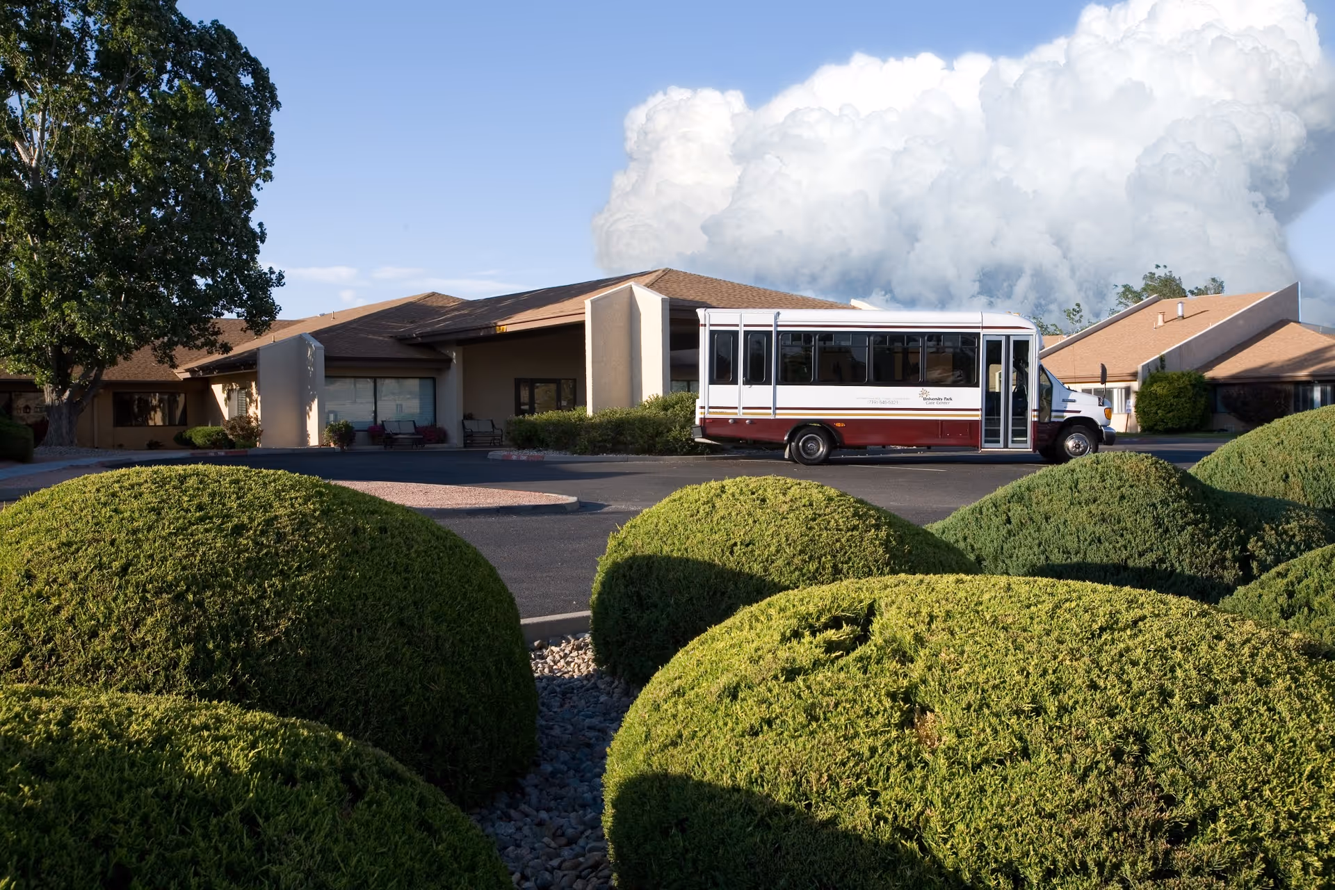 Front exterior of a single-story care center with rounded shrubs and a parked shuttle bus under a cloudy sky.