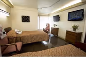A well-lit senior living room with two beds, dressers, a wall TV, and a window.