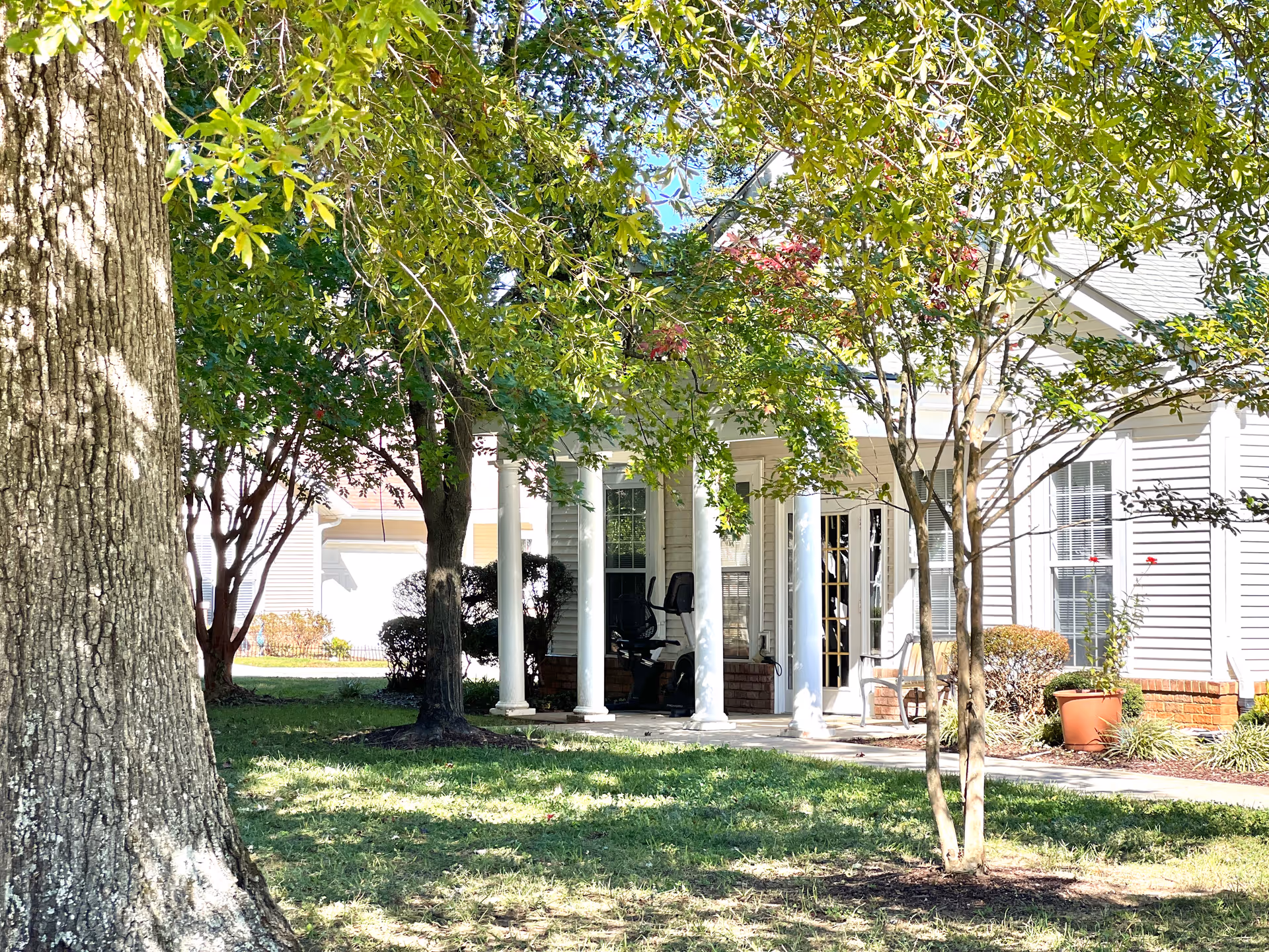 Porch and front facade of a single-story retirement community building with white columns, trees, lawn, and potted plants.