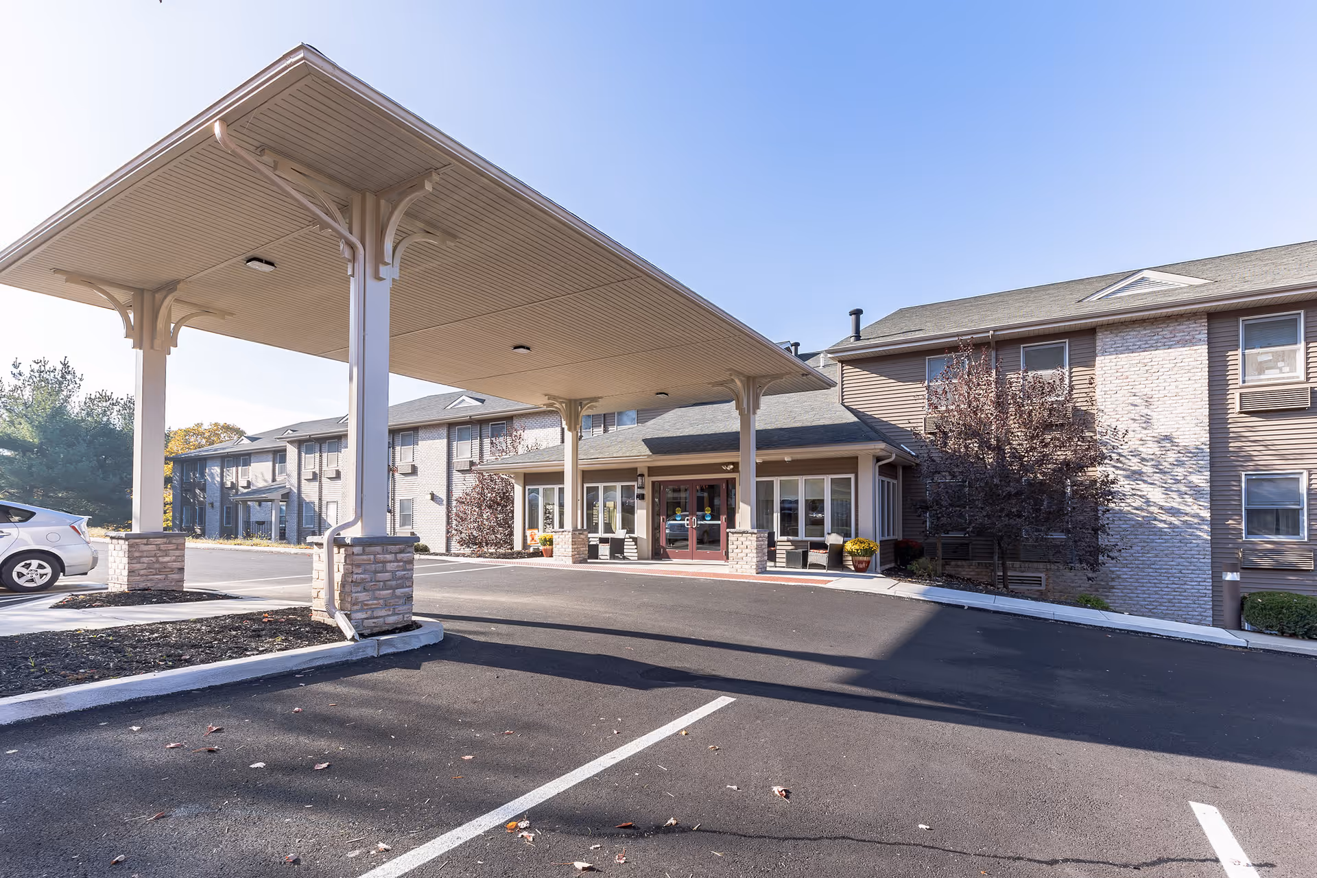 Front entrance of a senior living facility with a covered porte-cochere, parking area, and a two-story brick-and-siding building.