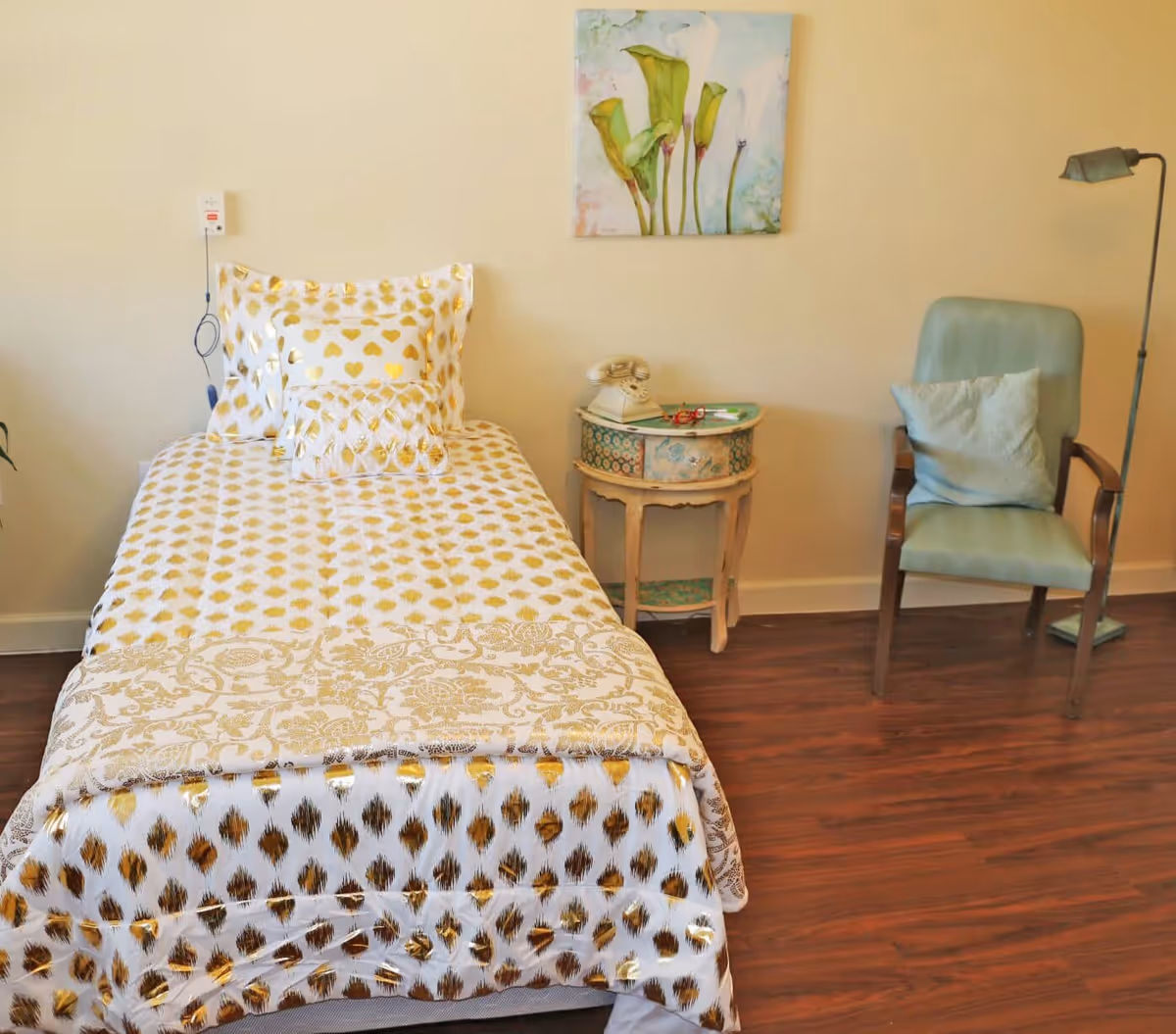 A neatly made single bed with white and gold patterned bedding in a room with wooden flooring. Next to the bed is a small round side table with a vintage telephone and some items on it. A light blue cushioned chair with a pillow and a tall floor lamp are positioned against the wall. Above the side table hangs a painting of green calla lilies on a light background.