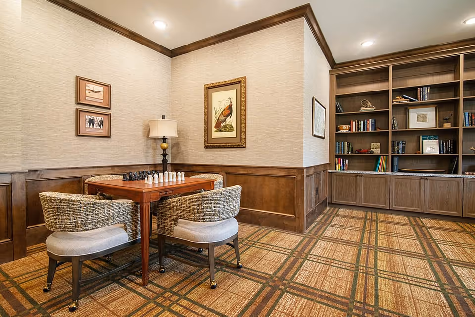 A cozy room with a wooden chess table and four cushioned chairs. The table has a chess set arranged on it. The room features wood paneling on the lower walls, beige wallpaper, framed artwork, a floor lamp, and a large built-in bookshelf filled with books and decorative items. The carpet has a plaid pattern in earth tones.