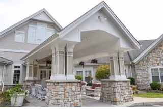 Exterior view of a senior living facility entrance with a covered porch supported by stone pillars, outdoor seating, and well-maintained landscaping.