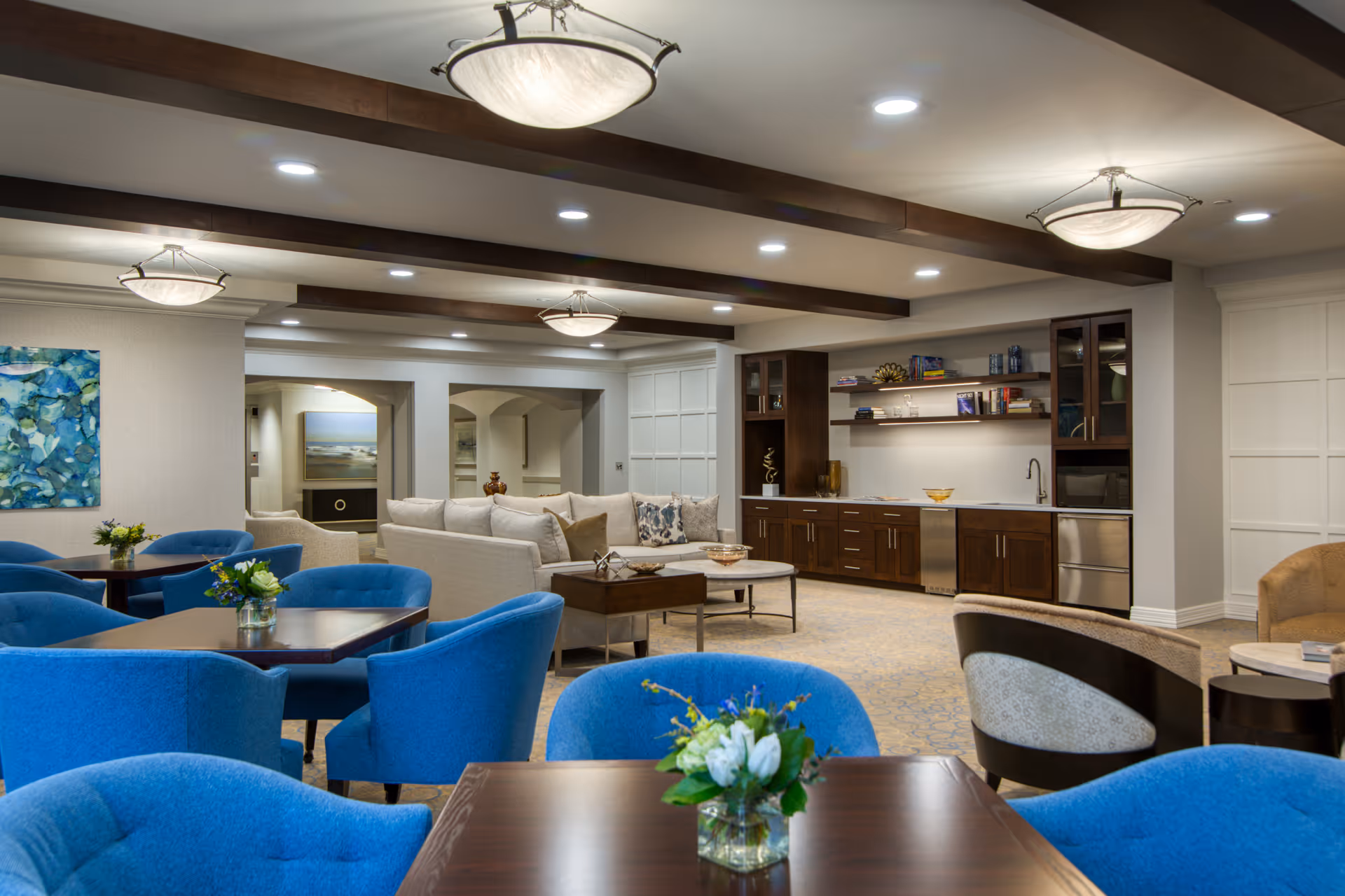 Well-lit senior living common room with blue dining chairs, tables, sofa seating, and a kitchenette area.