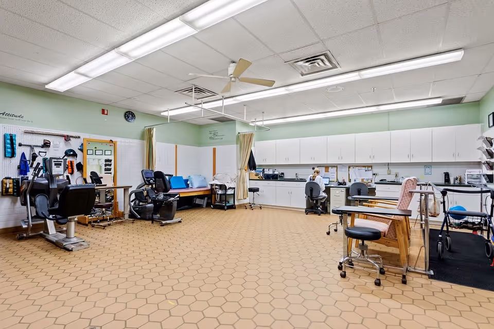 Spacious rehabilitation therapy room with exercise machines, treatment chairs and tables, cabinets, and a staff desk.