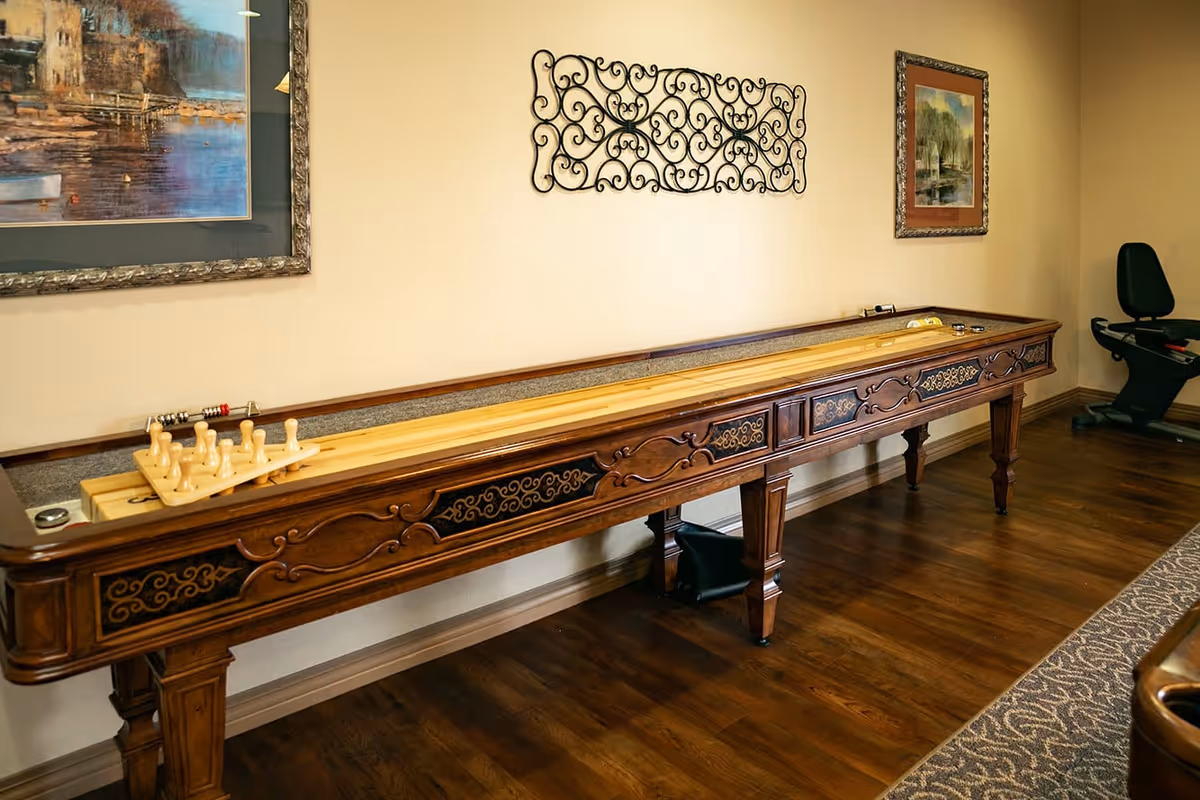 A long wooden shuffleboard table with decorative carvings is placed against a beige wall. On the table, there are shuffleboard pucks and a wooden rack holding additional pucks. The wall above the table features two framed paintings and a decorative wrought iron wall art piece. The floor is wooden, and there is a patterned carpet partially visible on the right side. A black exercise bike is positioned in the corner of the room.