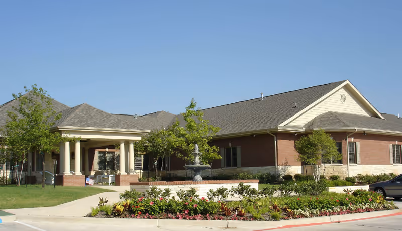 Exterior view of a single-story brick building with a covered entrance supported by columns, surrounded by landscaped greenery and a central water fountain under a clear blue sky.