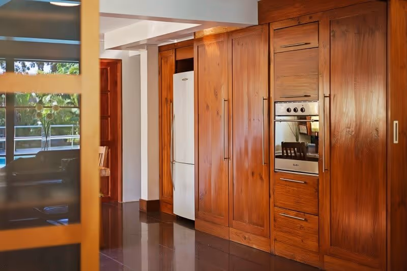 A kitchen area featuring wooden cabinets with built-in appliances including an oven and a refrigerator. There is a glimpse of a dining area with a chair and a window showing greenery outside.