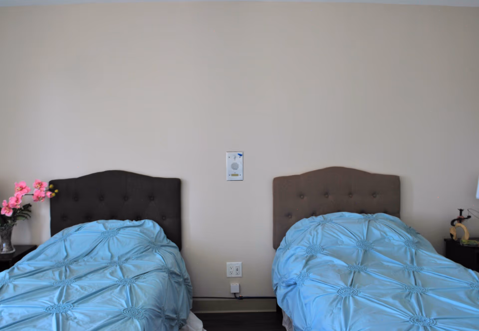 Two twin beds with tufted headboards and light blue comforters in a bedroom. There is a small nightstand with a vase of pink flowers on the left side and another nightstand with a lamp and decorative item on the right side. The wall behind the beds is plain with a small intercom or speaker device mounted between the beds.