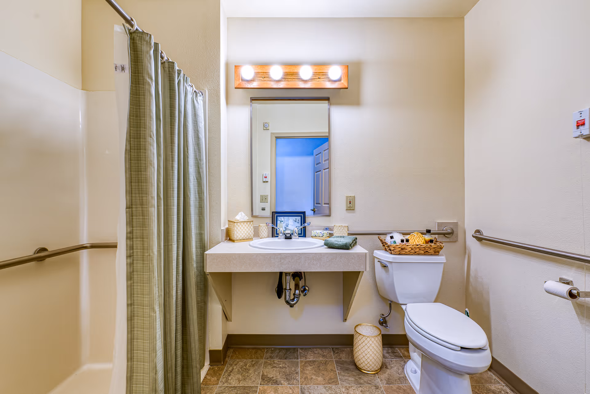 A clean and accessible bathroom featuring a shower with a green plaid curtain, a wall-mounted sink with a mirror and four lights above it, a toilet with a basket of toiletries on top, and grab bars along the walls for support.