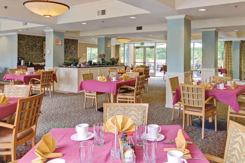 A bright dining room with multiple tables covered in magenta tablecloths and set with yellow folded napkins, white cups, glasses, and silverware. The room has light blue walls with white wainscoting, carpeted floors, and large windows letting in natural light. There are wooden chairs around the tables and a central counter with plants and flowers.