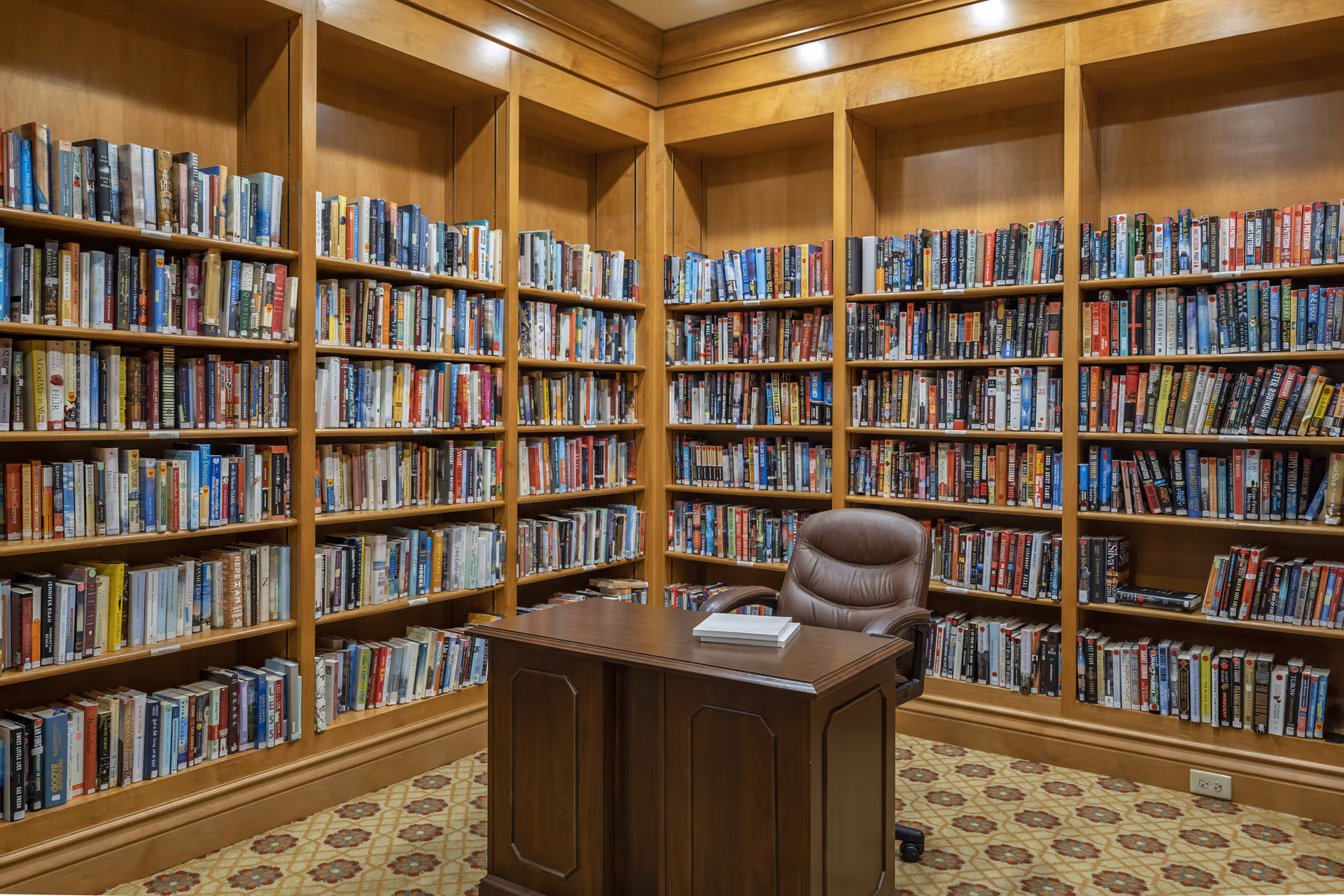 A cozy library room with wooden bookshelves filled with numerous books lining the walls. In the center, there is a wooden desk with a few books on top and a brown leather office chair behind it. The floor has a patterned carpet.