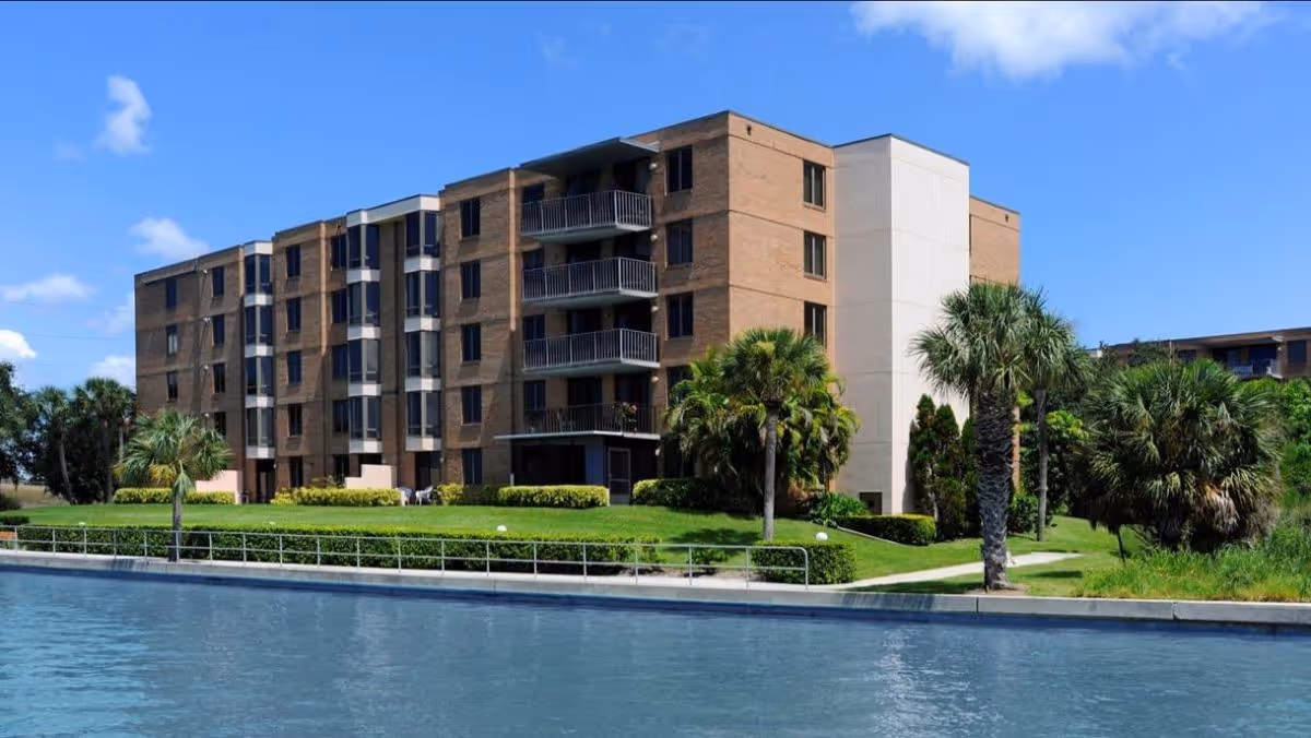 Exterior view of a multi-story senior living facility building with balconies, surrounded by green lawns, palm trees, and a body of water in the foreground under a blue sky with a few clouds.