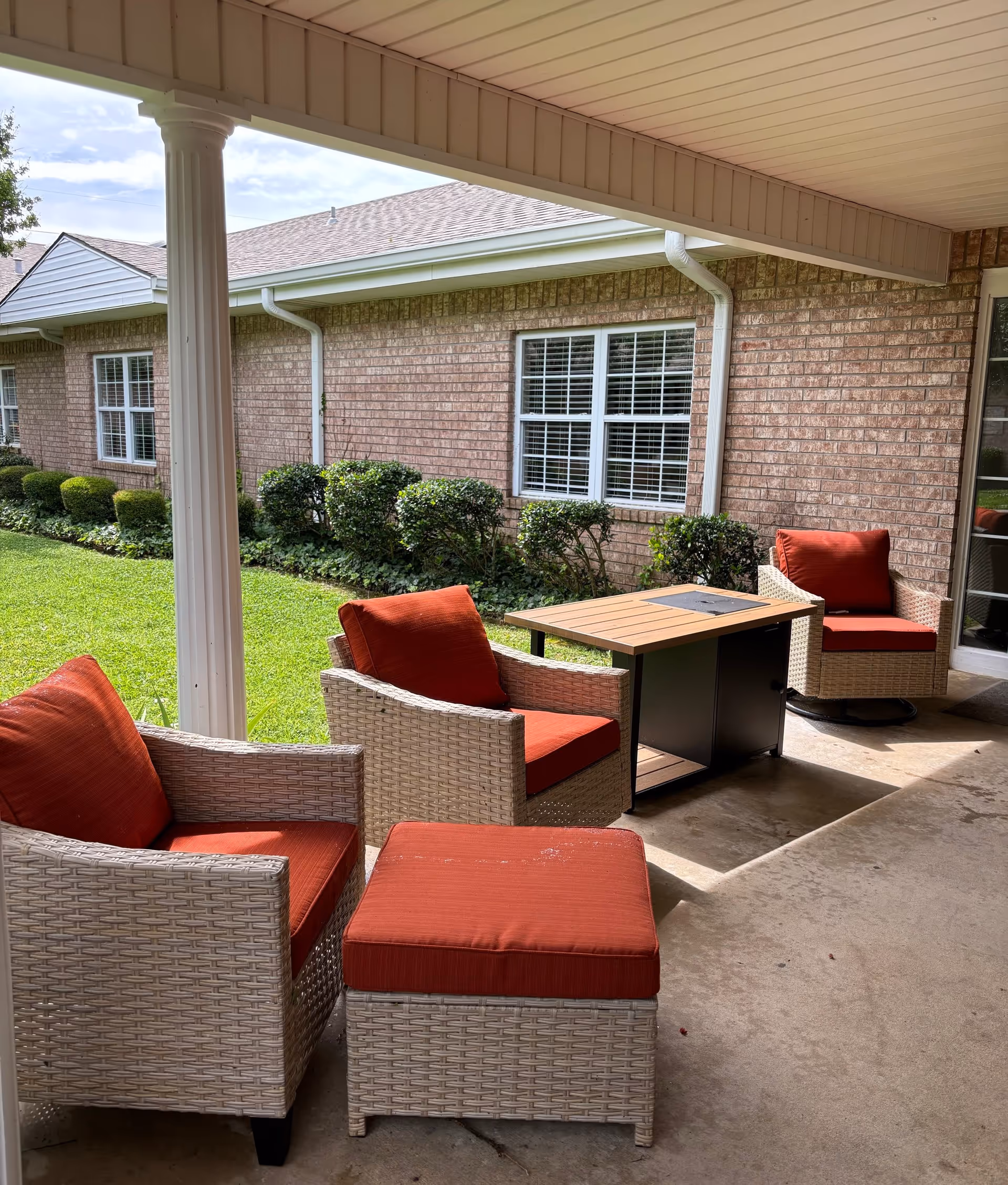 Covered outdoor patio area with three wicker chairs and a matching ottoman, all with red cushions, arranged around a rectangular wooden table. The patio overlooks a well-maintained lawn and shrubbery in front of a brick building with white-framed windows.