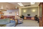 A spacious senior living common area with comfortable seating including a green sofa and a patterned armchair. There is a wooden side table between the seats. In the background, there is a long desk with two office chairs and a plant on top. The walls are painted in neutral tones with two wall-mounted lights above the desk. To the left, there is a dining area with tables and chairs, and a kitchen area is partially visible. The room is well-lit with ceiling lights and has carpeted flooring.