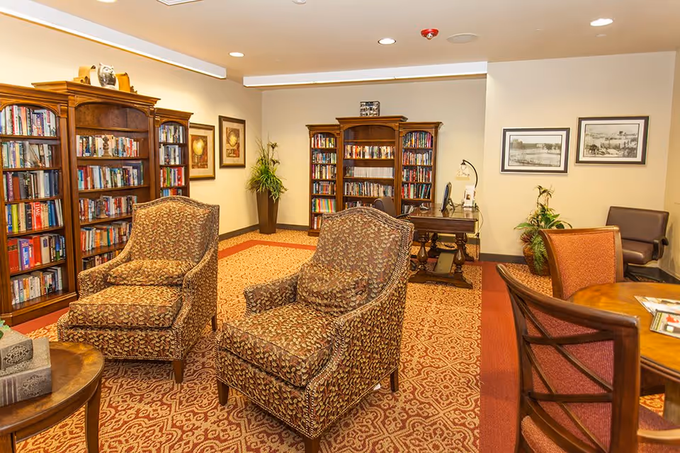 A cozy library room in an assisted living facility featuring two patterned armchairs in the foreground, wooden bookshelves filled with books along the walls, a wooden desk with a computer, framed artwork on the walls, and a round table with chairs on the right side. The room has warm lighting and a patterned carpet.