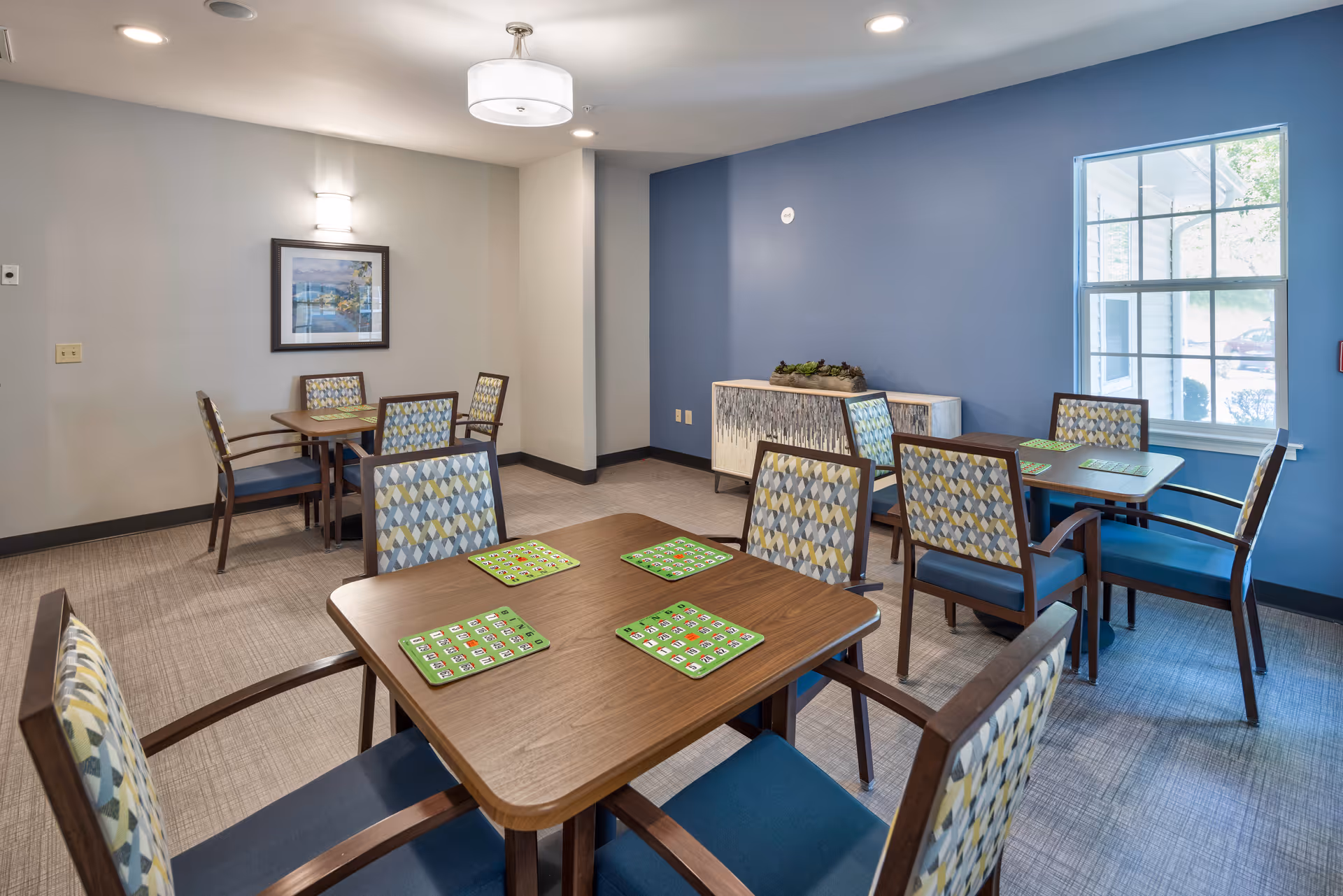 A bright and clean common room with multiple square wooden tables and patterned chairs arranged around them. Each table has green bingo cards placed on top. The room has a blue accent wall with a decorative sideboard and a window letting in natural light. A framed picture hangs on a light gray wall, and ceiling lights illuminate the space.