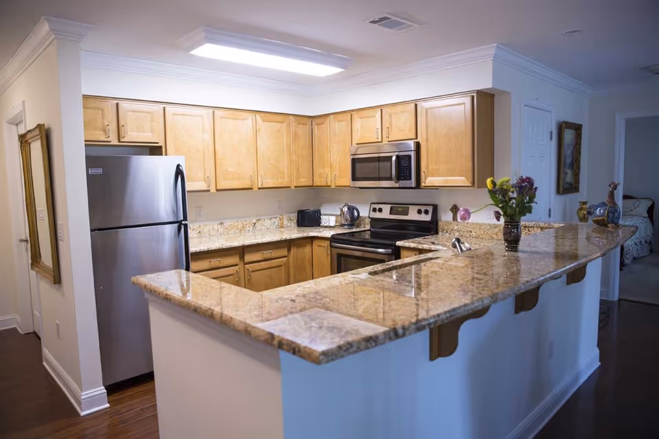 A modern kitchen with light wood cabinets, granite countertops, and stainless steel appliances including a refrigerator, microwave, and stove. There is a vase with flowers on the countertop and a toaster and kettle on the counter near the stove. The kitchen has a bright overhead light and an open layout with a bar-style counter facing the living area.