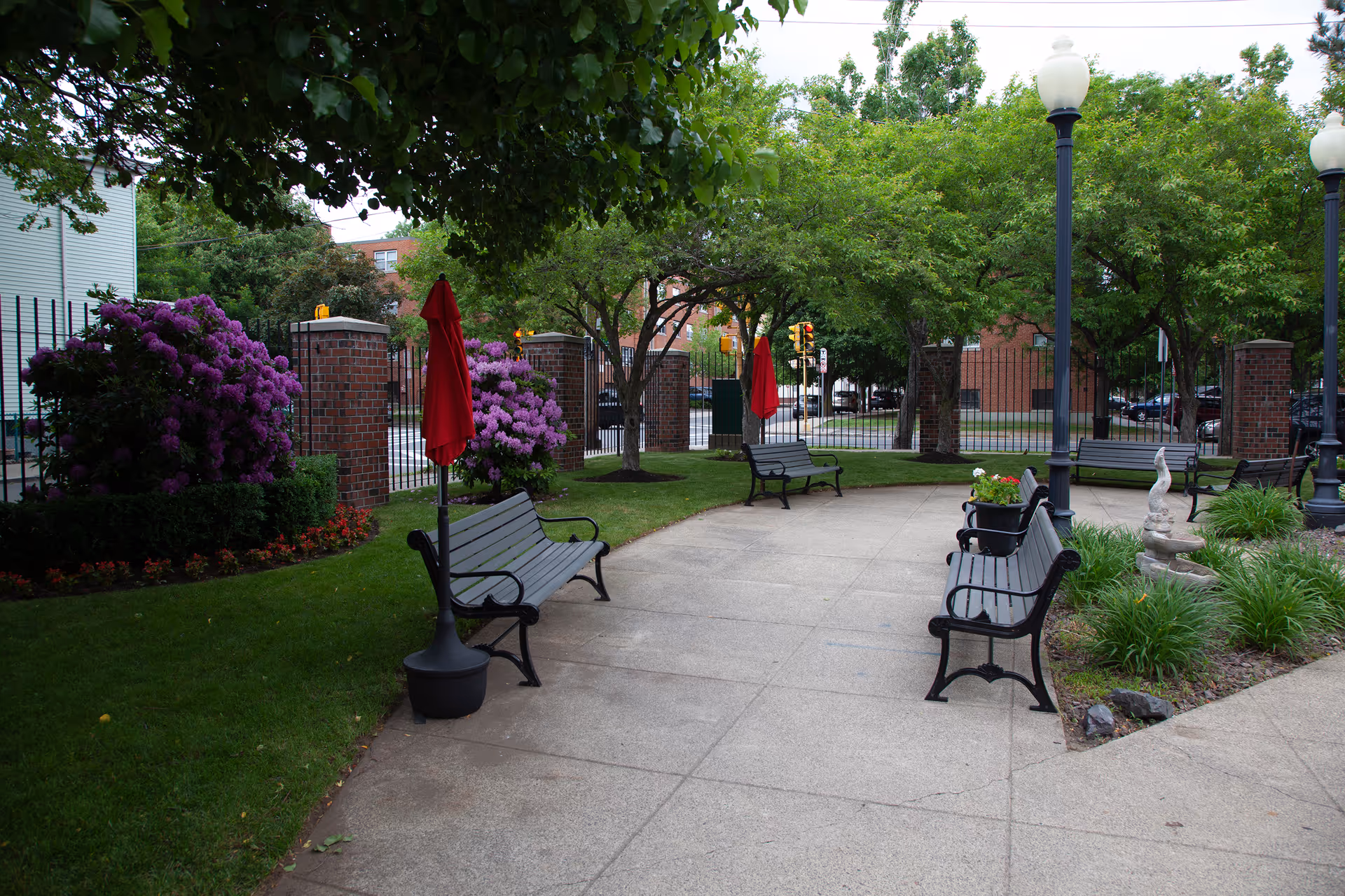 Outdoor courtyard with paved walkways, benches, lamp posts, red umbrellas, and flowering shrubs.