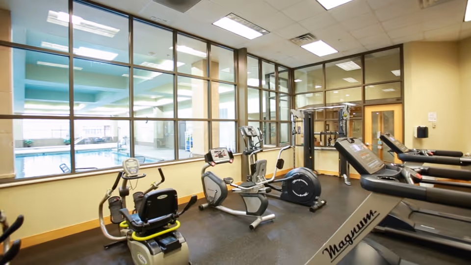 Interior fitness room with treadmills, recumbent bike and other exercise equipment facing windows that look out onto an indoor pool.