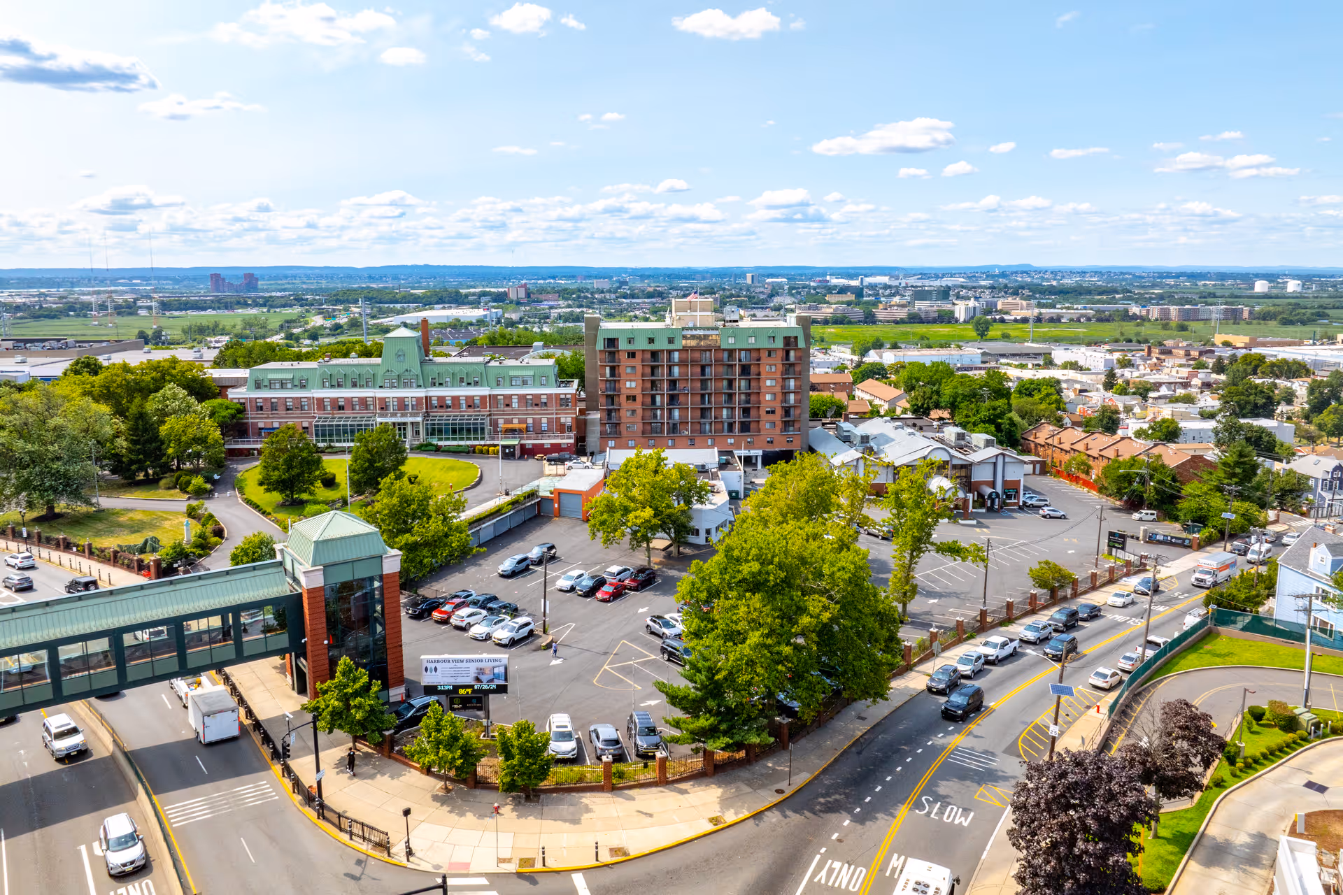Aerial view of Harbour View Senior Living Community showing multiple buildings with green roofs, surrounding parking lots, trees, and nearby roads with cars. The background includes a wide cityscape under a partly cloudy sky.