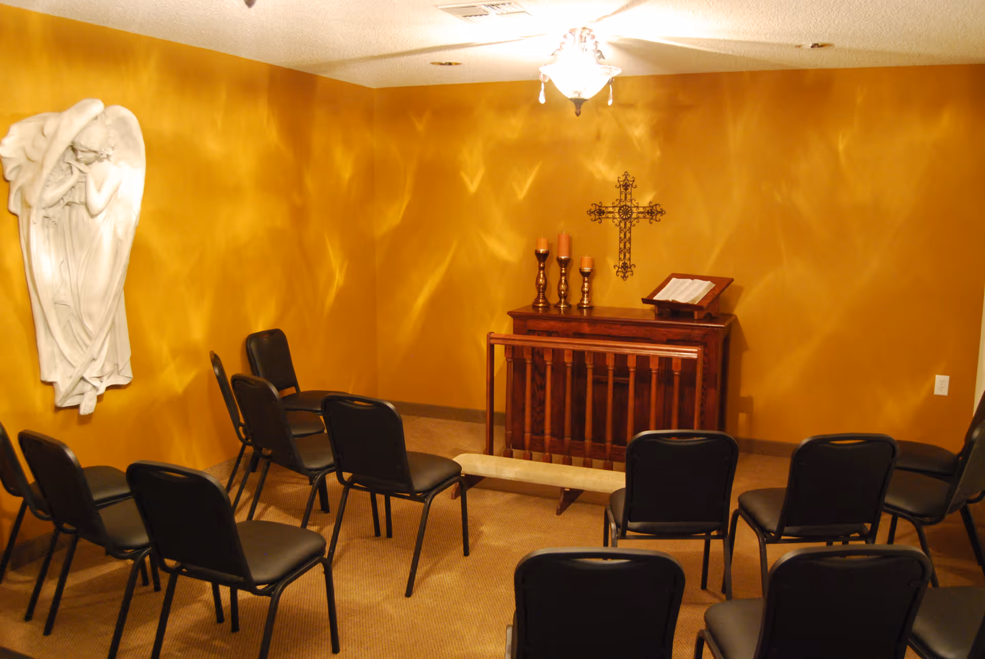 Small chapel-style room with rows of chairs facing a wooden altar, a wall cross, candles, and an angel relief on a golden wall.