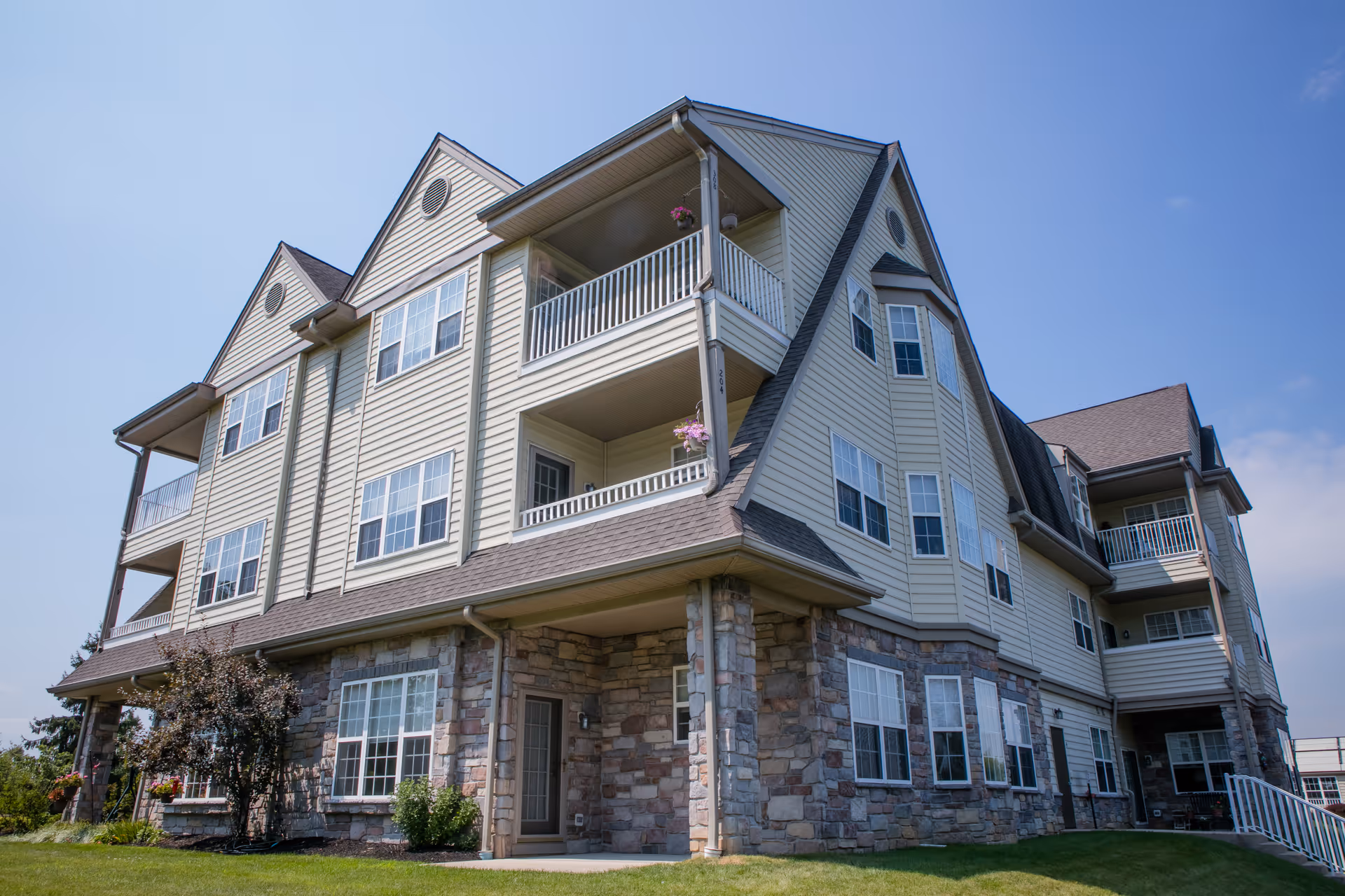 Exterior view of a multi-story senior living facility building with beige siding and stone accents, featuring multiple windows and balconies under a clear blue sky.