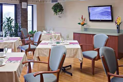 Morris Hills Center dining room with tables set for meals, chairs, plants, and a wall-mounted TV.