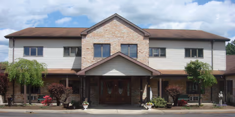 Front exterior view of a two-story building with a brick and siding facade, a covered entrance with double wooden doors, and landscaped shrubs and small trees on either side of the entrance under a partly cloudy sky.