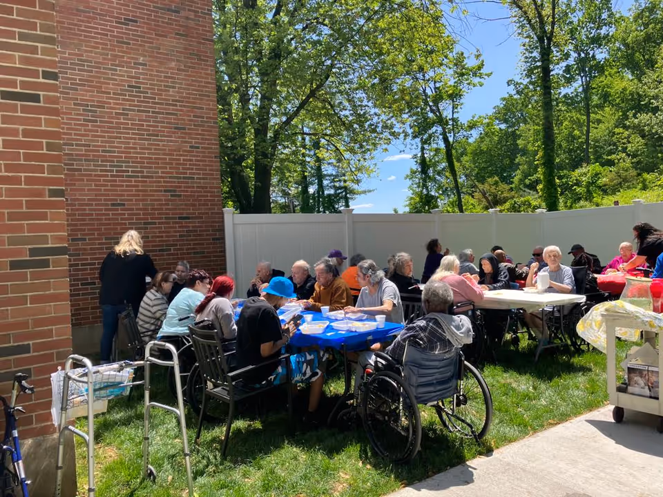 A group of elderly people sitting outdoors at tables covered with blue and white tablecloths, enjoying a meal together in a fenced grassy area next to a brick building. Some individuals are seated in wheelchairs, and there is a walker visible near the building. Trees and a clear blue sky are in the background.