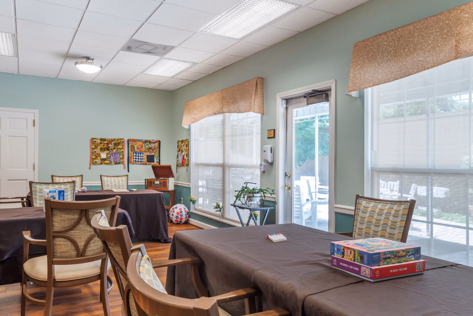 A well-lit activity room with several tables covered in dark brown tablecloths and chairs arranged around them. On one table, there are two puzzle boxes and some markers. The room has light green walls, large windows with beige valances, and a door leading outside to a patio area with white chairs. Decorative wall hangings and a vintage-style radio are visible on the far wall.