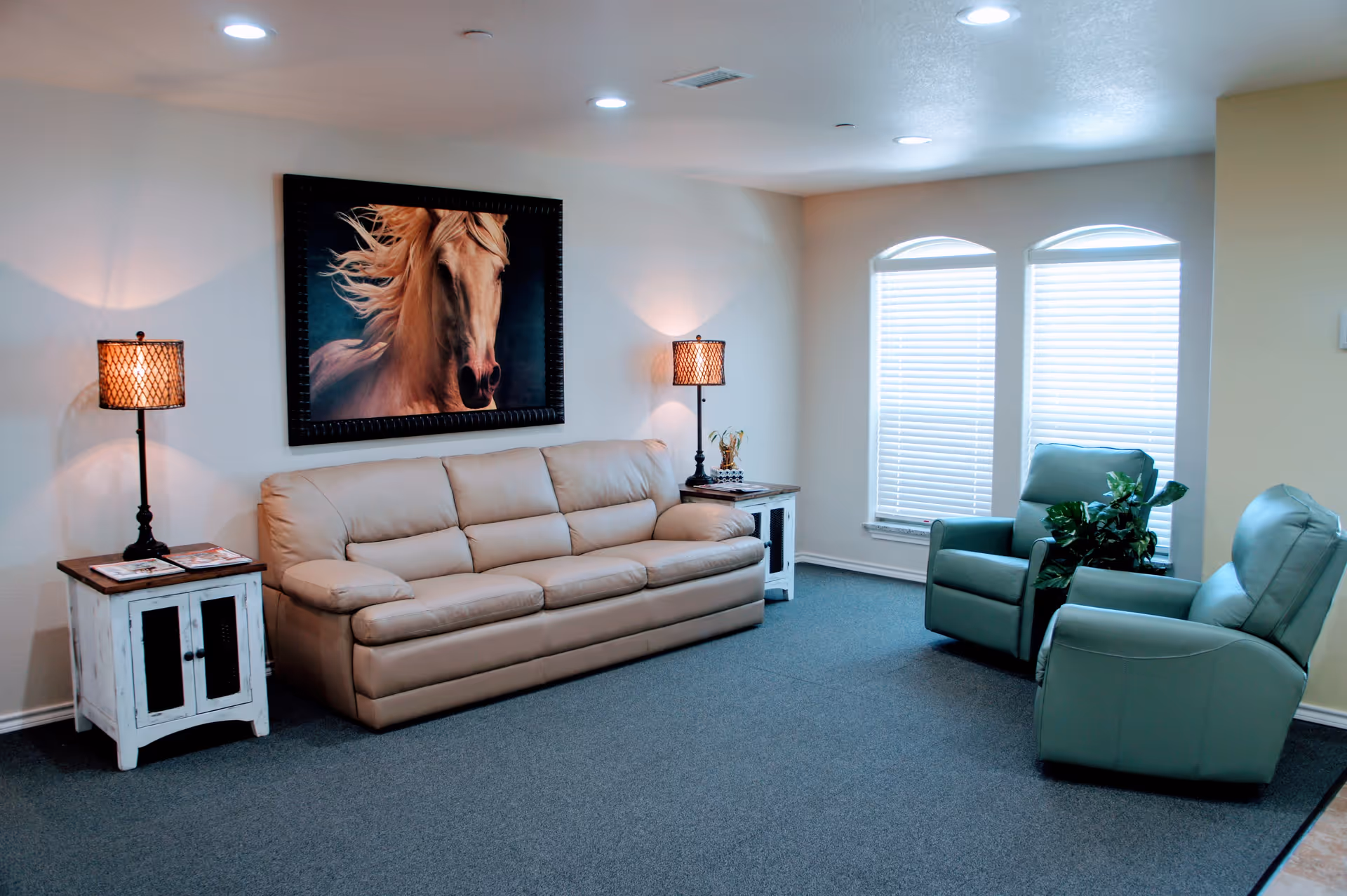 A cozy living room with a beige leather sofa, two green armchairs, two side tables with lamps, a large framed picture of a horse on the wall, and two windows with blinds letting in natural light.