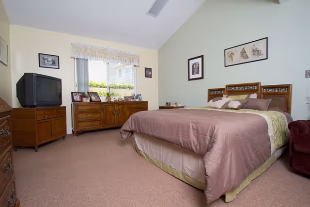 Neat bedroom with a large made bed, wooden dressers and a TV beneath a window with framed pictures on the walls.