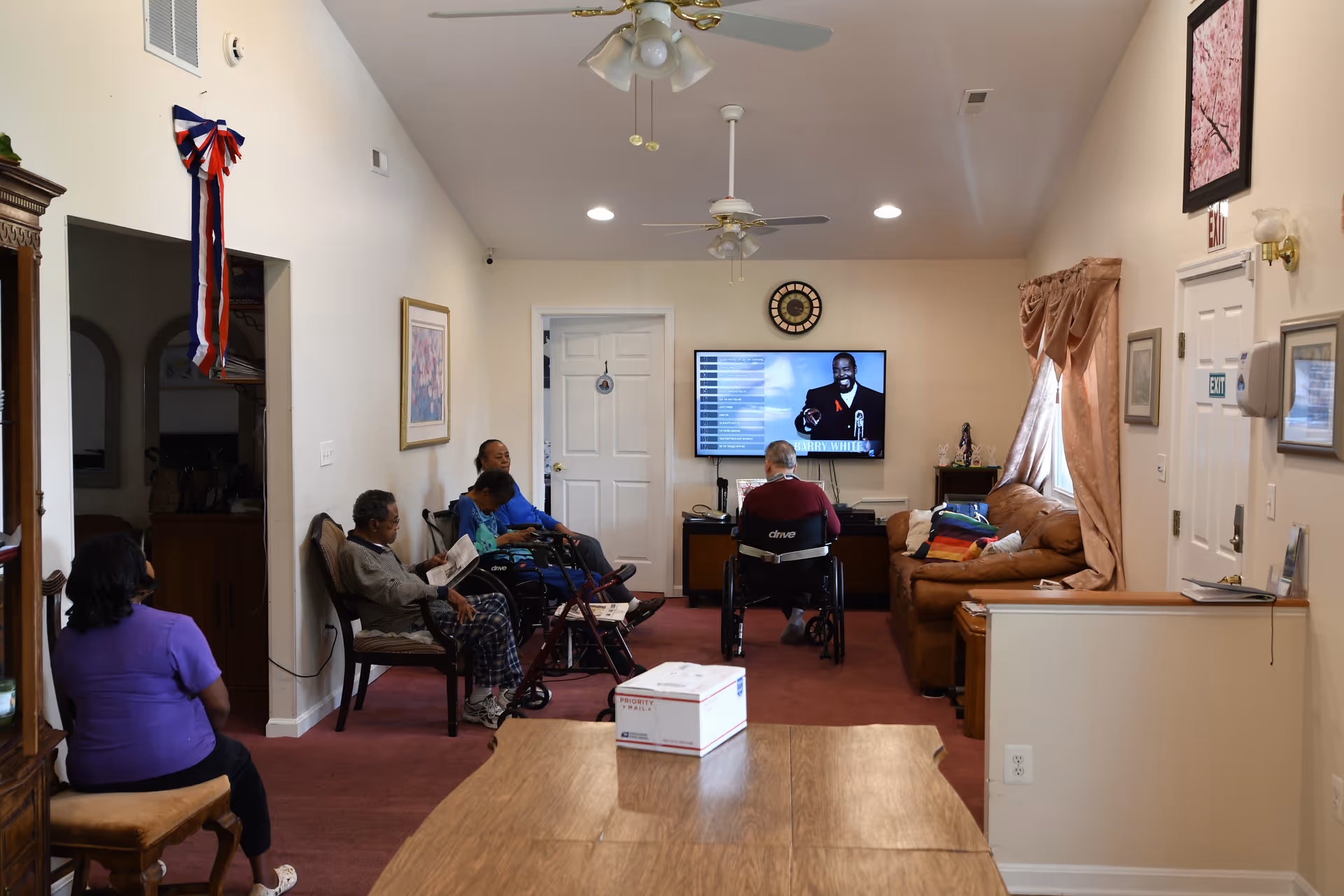 A group of elderly individuals sitting in a living room area of an assisted living facility, some in wheelchairs, watching a television mounted on the wall. The room has beige walls, ceiling fans, a brown couch with a colorful blanket, framed pictures, and a wooden table in the foreground.